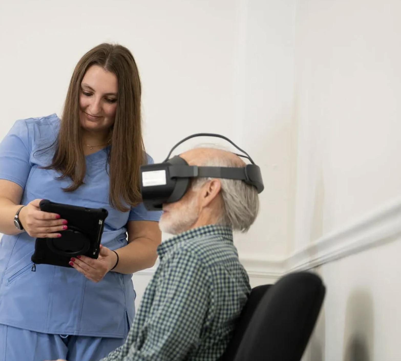 A nurse in blue scrubs shows a tablet to an older man wearing a virtual reality headset, seated on a chair in a white room.